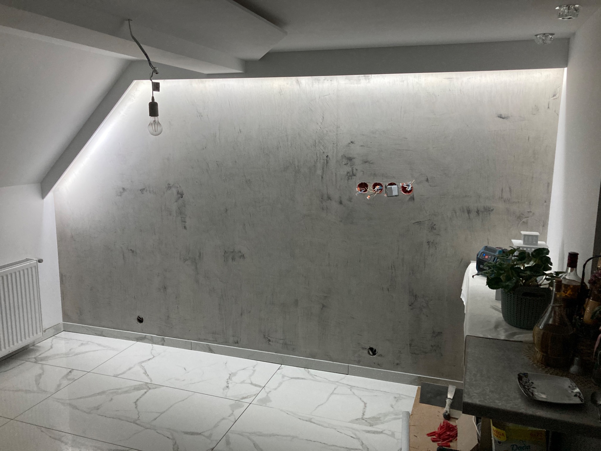 Ściana z efektem dekoracyjnym w szarościach, podświetlona listwą LED, widoczne gniazdka elektryczne bez osprzętu, białe płytki podłogowe imitujące marmur, surowa żarówka na kablu