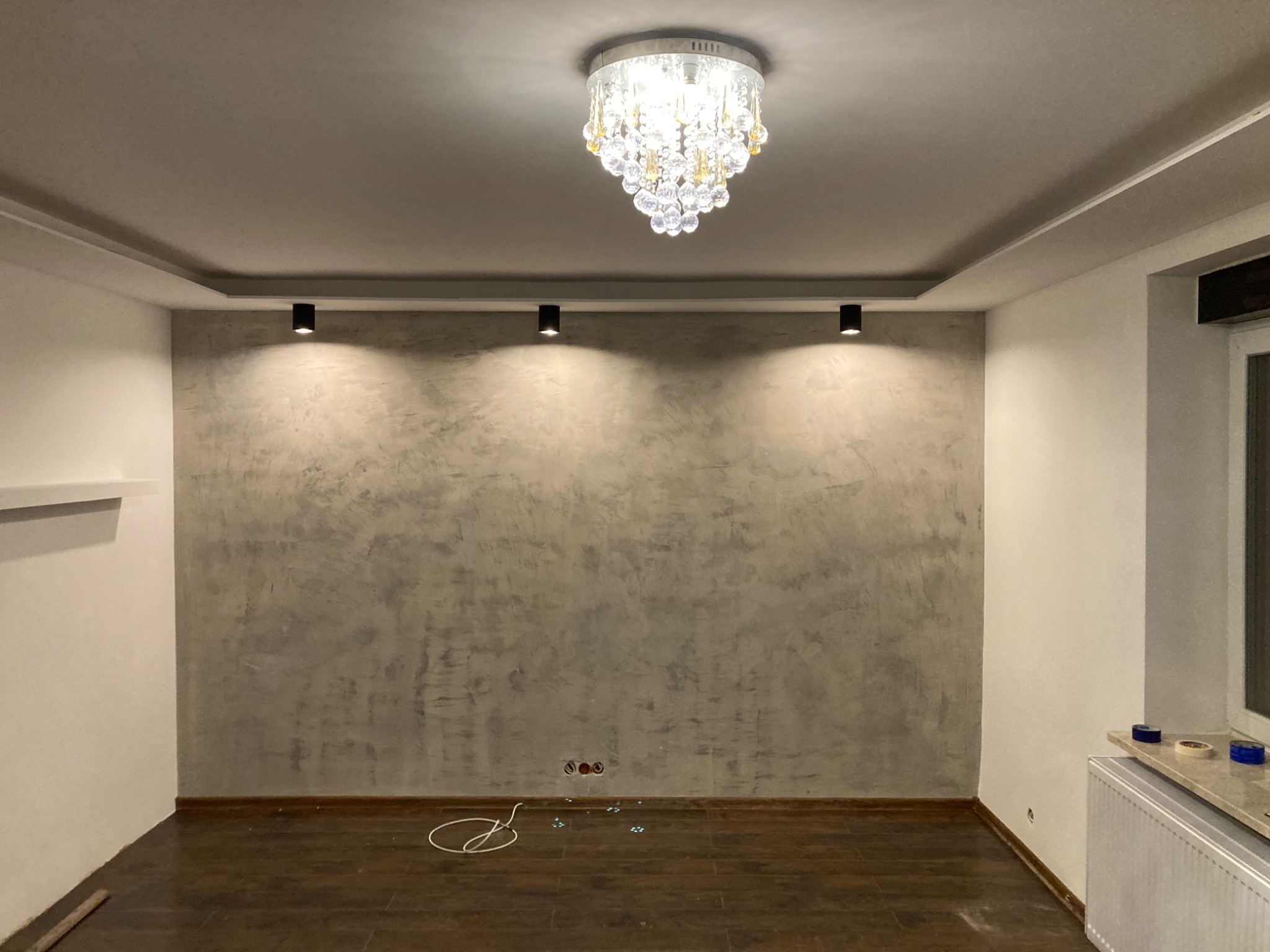 Pokój po remoncie: ściana pokryta szarą, dekoracyjną gładzią, na suficie kryształowy żyrandol i trzy czarne reflektory punktowe, brązowe panele podłogowe, widoczne gniazdka elektryczne bez osprzętu.