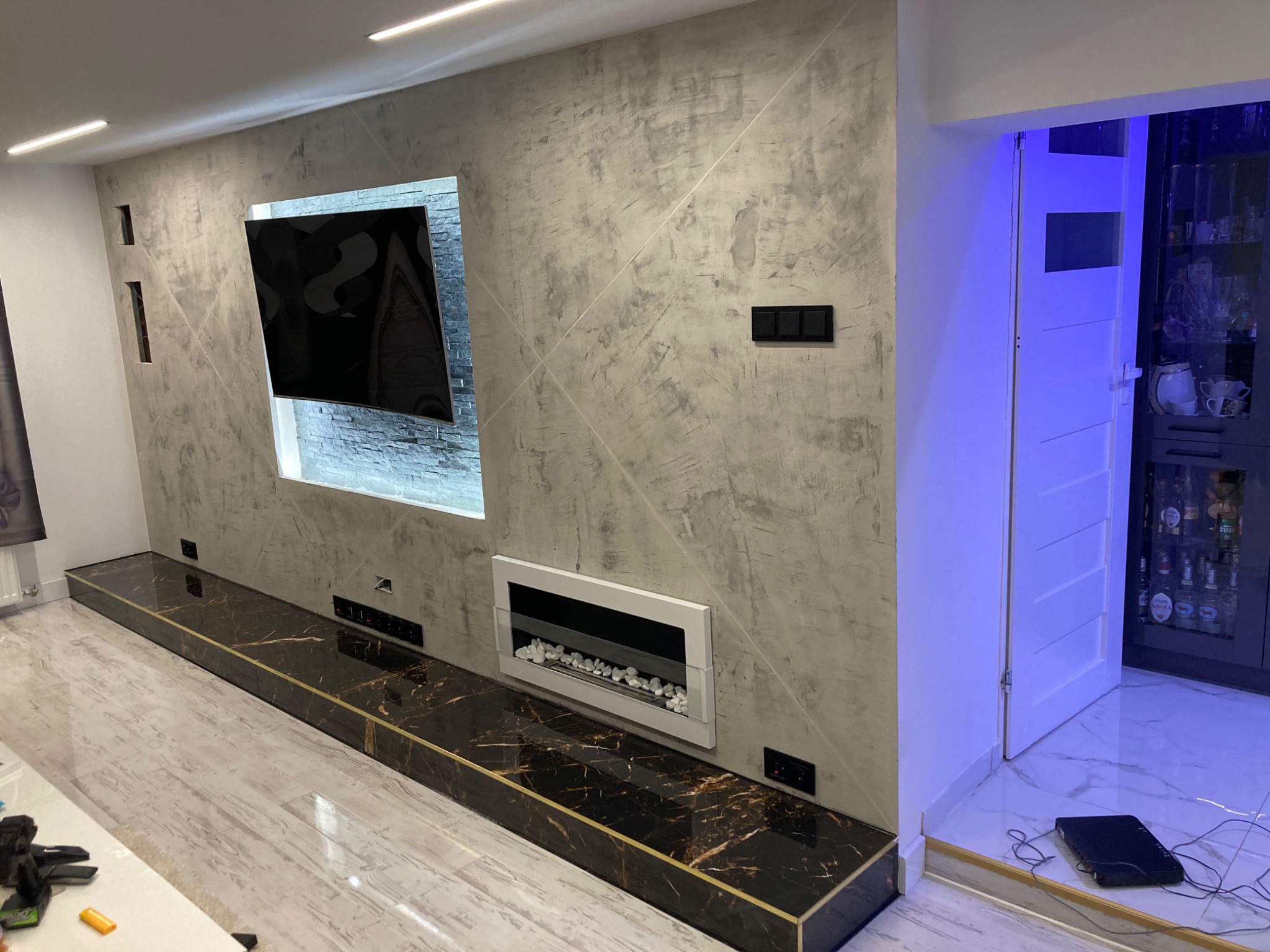 Nowoczesny salon z szarą ścianą o fakturze betonu, wnęką na telewizor z kamienną okładziną i elektrycznym kominkiem z białymi kamykami, podświetlany sufit, marmurowy blat z złotymi akcentami...