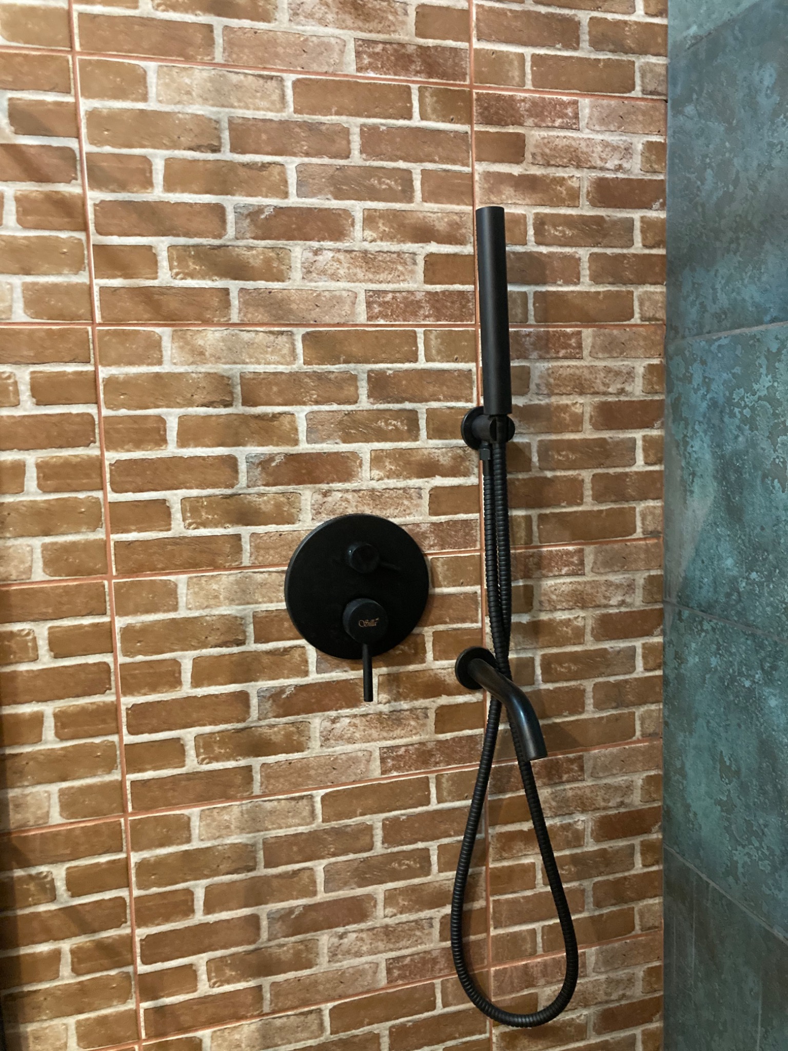 Ciemna, matowa armatura prysznicowa na ścianie wyłożonej brązowymi płytkami imitującymi cegłę, z elementami dekoracyjnymi w odcieniach szarości.
