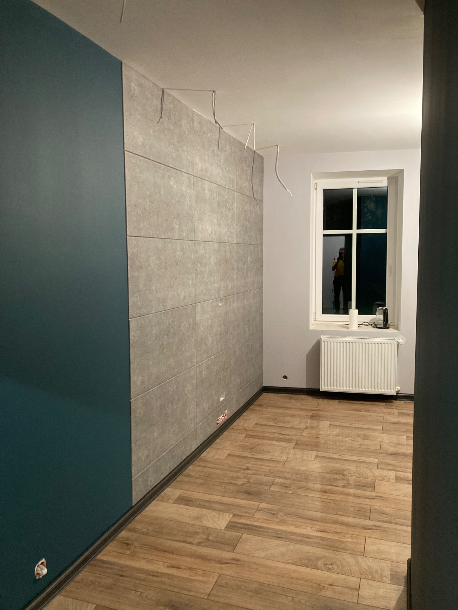 Wnętrze pokoju po częściowym remoncie: pomalowana na ciemnozielono ściana kontrastuje z szarą ścianą wyłożoną panelami imitującymi beton, widoczne kable wystające ze ściany, drewnopodobna podłoga...