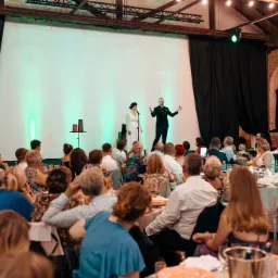 Iluzjonista w czarnym garniturze prezentuje trik z czerwonymi kulkami na scenie podczas wesela, obok stoi kobieta w białej sukni, widownia siedzi przy okrągłych stołach.