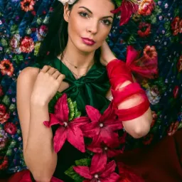 Portret kobiety z wiankiem z białych i żółtych róż na głowie, ubrana w zieloną sukienkę ozdobioną czerwonymi kwiatami z perłami i czerwonymi wstążkami na ramieniu, na tle kwiecistej tapety.
