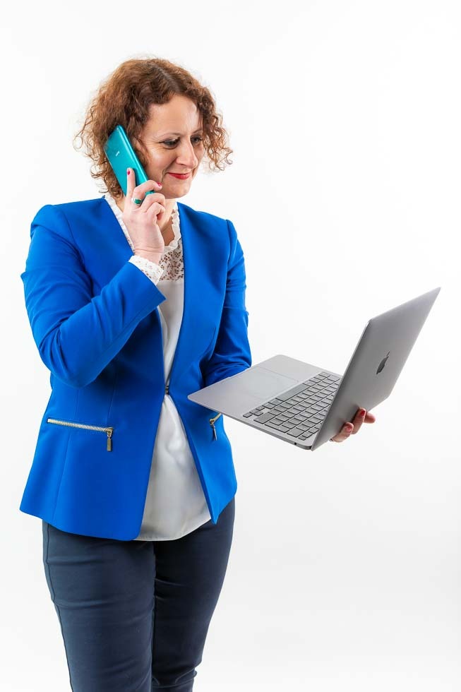 Kobieta w niebieskiej marynarce rozmawia przez telefon i trzyma otwarty laptop na białym tle.