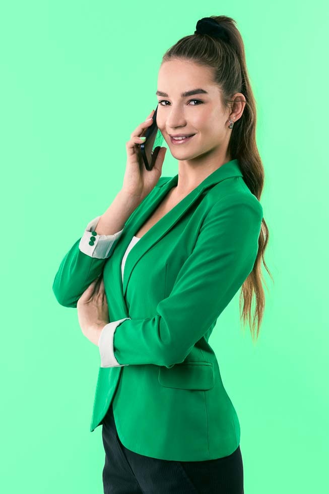 Uśmiechnięta kobieta w zielonej marynarce rozmawia przez telefon na jednolitym, jasnym, miętowym tle.