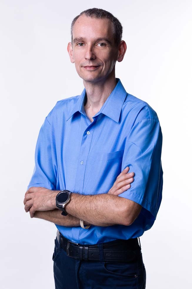 Portret mężczyzny w błękitnej koszuli, z założonymi rękami, na białym tle. Widoczny zegarek na lewym nadgarstku i skórzany pasek przy spodniach.