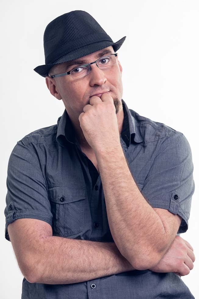 Mężczyzna w czarnym kapeluszu i okularach, z brodą, w szarej koszuli, zamyślony, z ręką przy ustach, na białym tle.