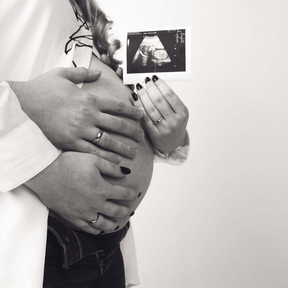 Czarno-białe zdjęcie dłoni kobiety z czarnym manicure i pierścionkiem zaręczynowym, trzymającej zdjęcie USG płodu na tle ciążowego brzucha, obejmowanego dłońmi mężczyzny z obrączką.