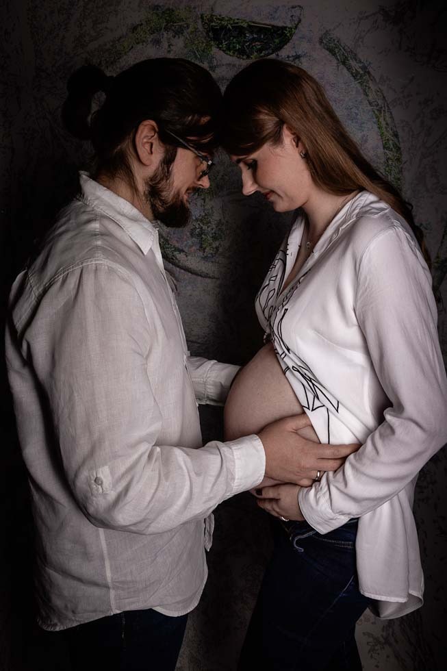 Przyszli rodzice w białych koszulach, dotykający ciążowego brzucha na tle ściany z wzorem przypominającym korę drzewa.