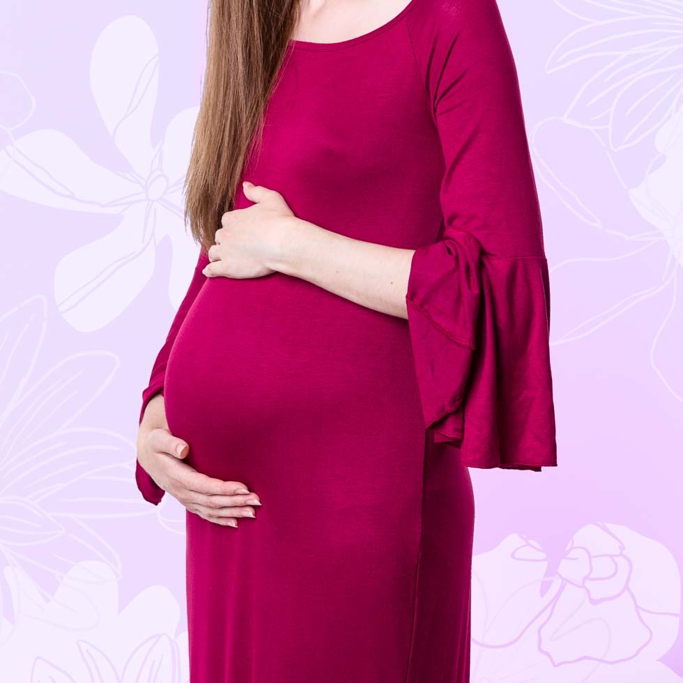 Kobieta w bordowej sukience z rękawami typu dzwonek, delikatnie obejmująca ciążowy brzuszek na tle z motywem kwiatowym.