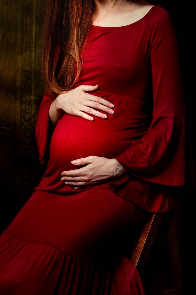 Kobieta w czerwonej sukni ciążowej siedzi, trzymając dłonie na brzuchu, z rudymi włosami opadającymi na ramię, na tle ciemnej, teksturowanej ściany.