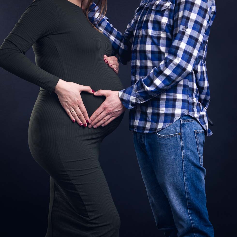 Przyszła mama w dopasowanej, ciemnozielonej sukience, z dłońmi ułożonymi w kształt serca na brzuchu, obok partner w niebieskiej koszuli w kratę i jeansach, na ciemnym tle.
