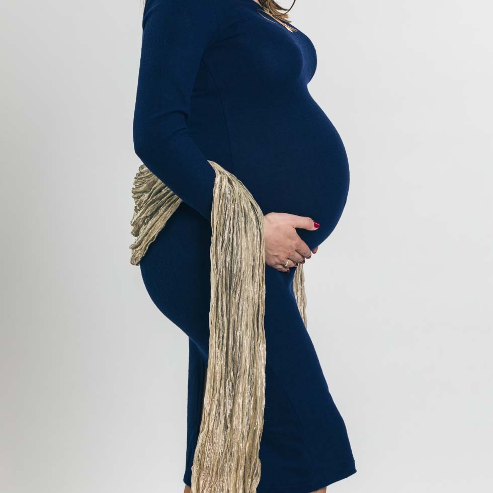 Kobieta w granatowej sukience ciążowej, z ręką na brzuchu i złotym szalem z frędzlami, na jasnym tle.