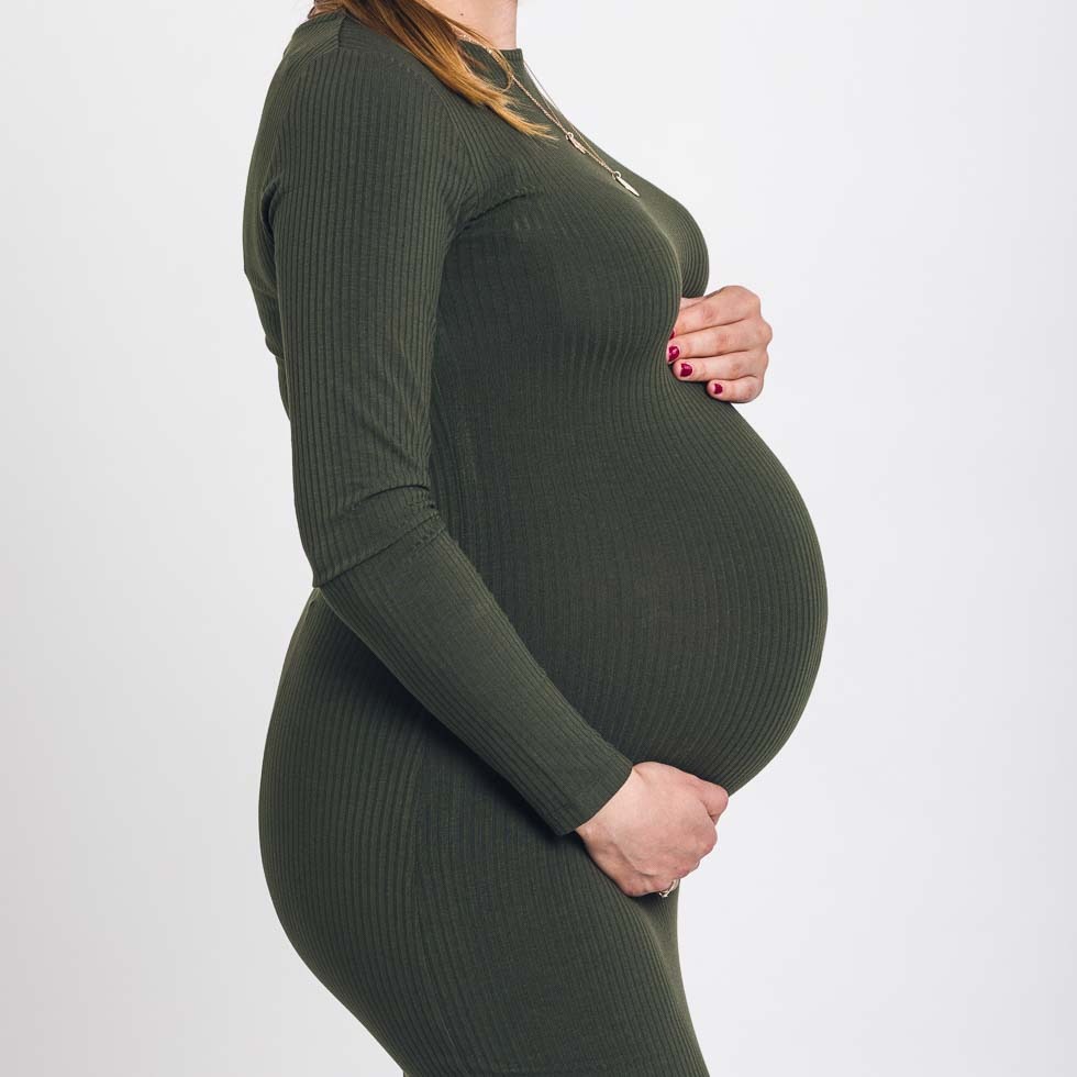 Kobieta w dopasowanej sukience w kolorze oliwkowym, trzymająca dłonie na dużym ciążowym brzuchu, widok z profilu na jasnym tle.