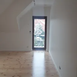 Puste poddasze po remoncie z jasną drewnianą podłogą, białymi ścianami z dekoracyjnymi naklejkami i oknem balkonowym z widokiem na zimowy ogród.