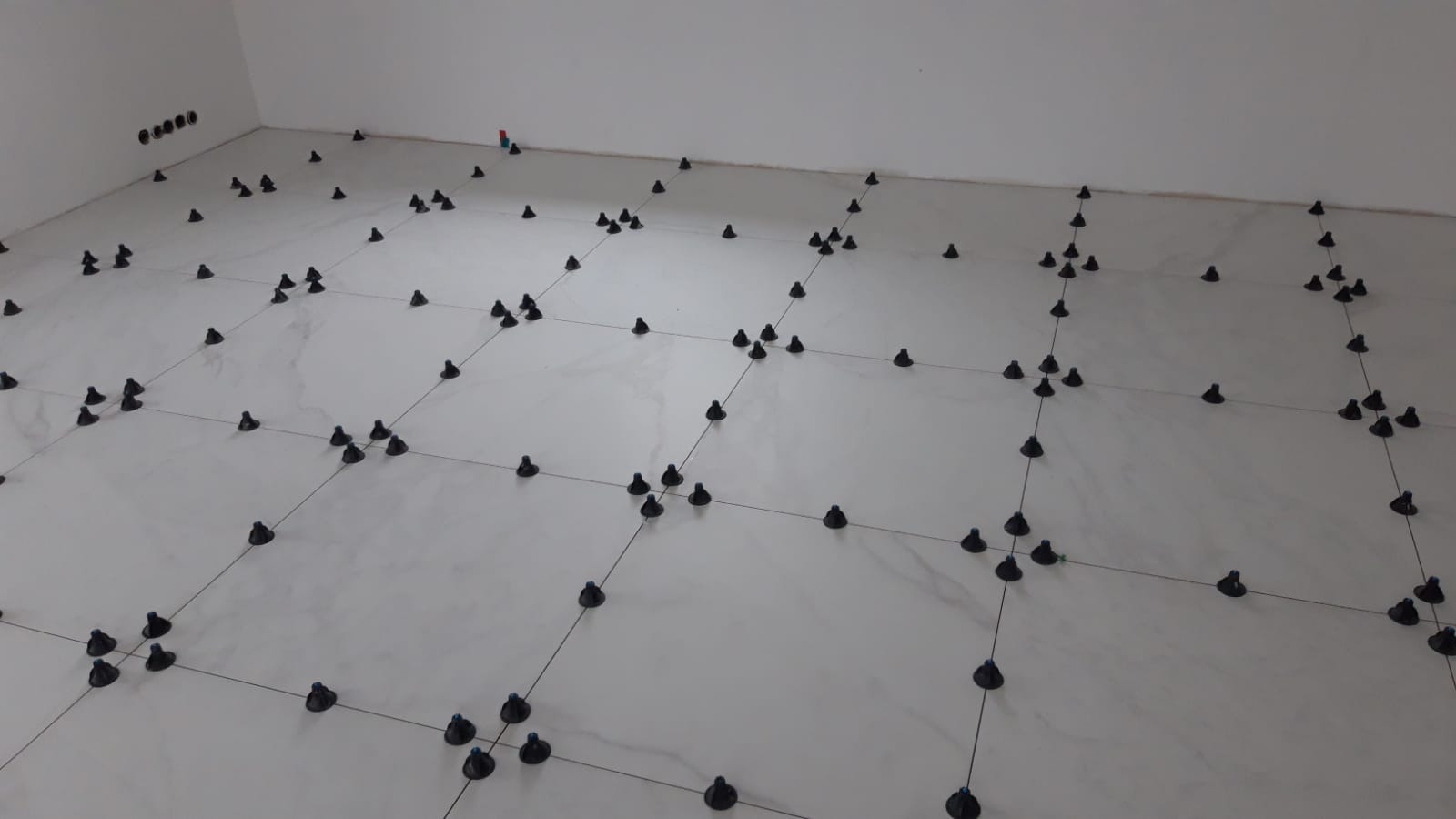 Białe płytki podłogowe w trakcie układania, z użyciem systemu poziomowania z czarnymi klipsami i sznurkami dystansowymi, widoczne gniazdka elektryczne w ścianie.