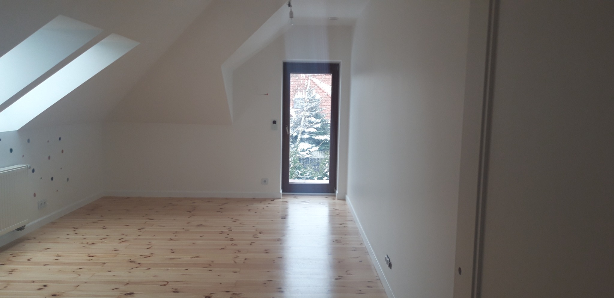 Puste poddasze po remoncie z jasną drewnianą podłogą, białymi ścianami z dekoracyjnymi naklejkami i oknem balkonowym z widokiem na zimowy ogród.