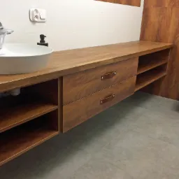 Nowoczesna łazienka z drewnianą szafką pod umywalką z szufladami z brązowymi skórzanymi uchwytami, białą okrągłą umywalką nablatową i toaletą w tle.