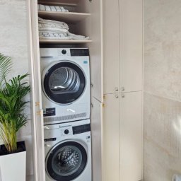 WOJCIECH DOMALEWSKI GARDER 4U - Biała szafa wnękowa z pralką i suszarką w łazience, z półkami na tekstylia. Minimalistyczny design, jasne wnętrze, roślina w doniczce obok.