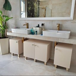 WOJCIECH DOMALEWSKI GARDER 4U - Nowoczesna łazienka z beżowymi szafkami pod umywalki, złotymi bateriami i nogami szafek. Umywalki prostokątne, białe. Na blacie akcesoria i roślina doniczkowa.