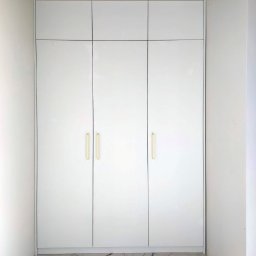 GARDER 4U - Minimalistyczna, biała zabudowa meblowa z połyskiem, trzema frontami i trzema górnymi szafkami, ze złotymi uchwytami, wkomponowana we wnękę, na tle marmurowej podłogi.