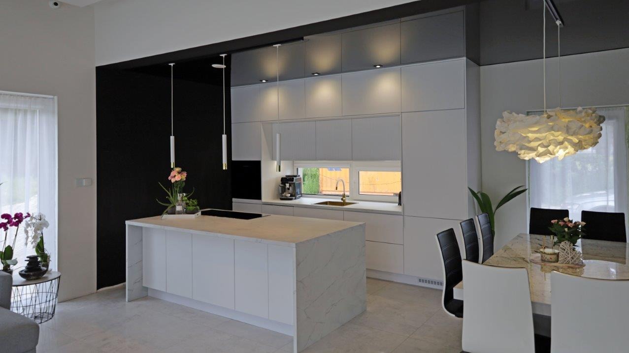 Nowoczesna kuchnia z wyspą i białymi frontami, minimalistyczne lampy wiszące, marmurowy blat wyspy, czarny akcent na ścianie, jasna podłoga i stół z krzesłami.