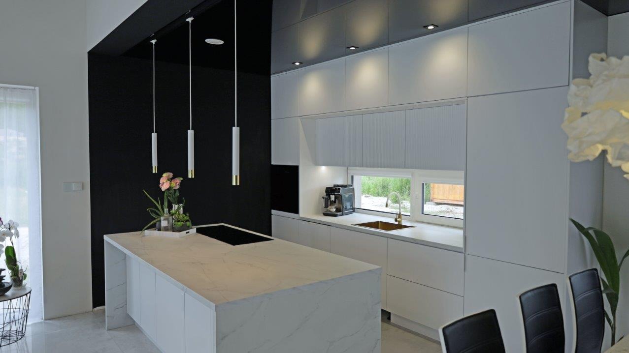 Nowoczesna, biała kuchnia z wyspą o marmurowym blacie, oświetlona wiszącymi lampami. Czarne akcenty i minimalistyczny design wnętrza.
