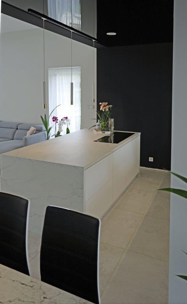 Nowoczesna kuchnia z wyspą o marmurowym blacie, minimalistyczne oświetlenie, czarna ściana w tle, widok z perspektywy jadalni.
