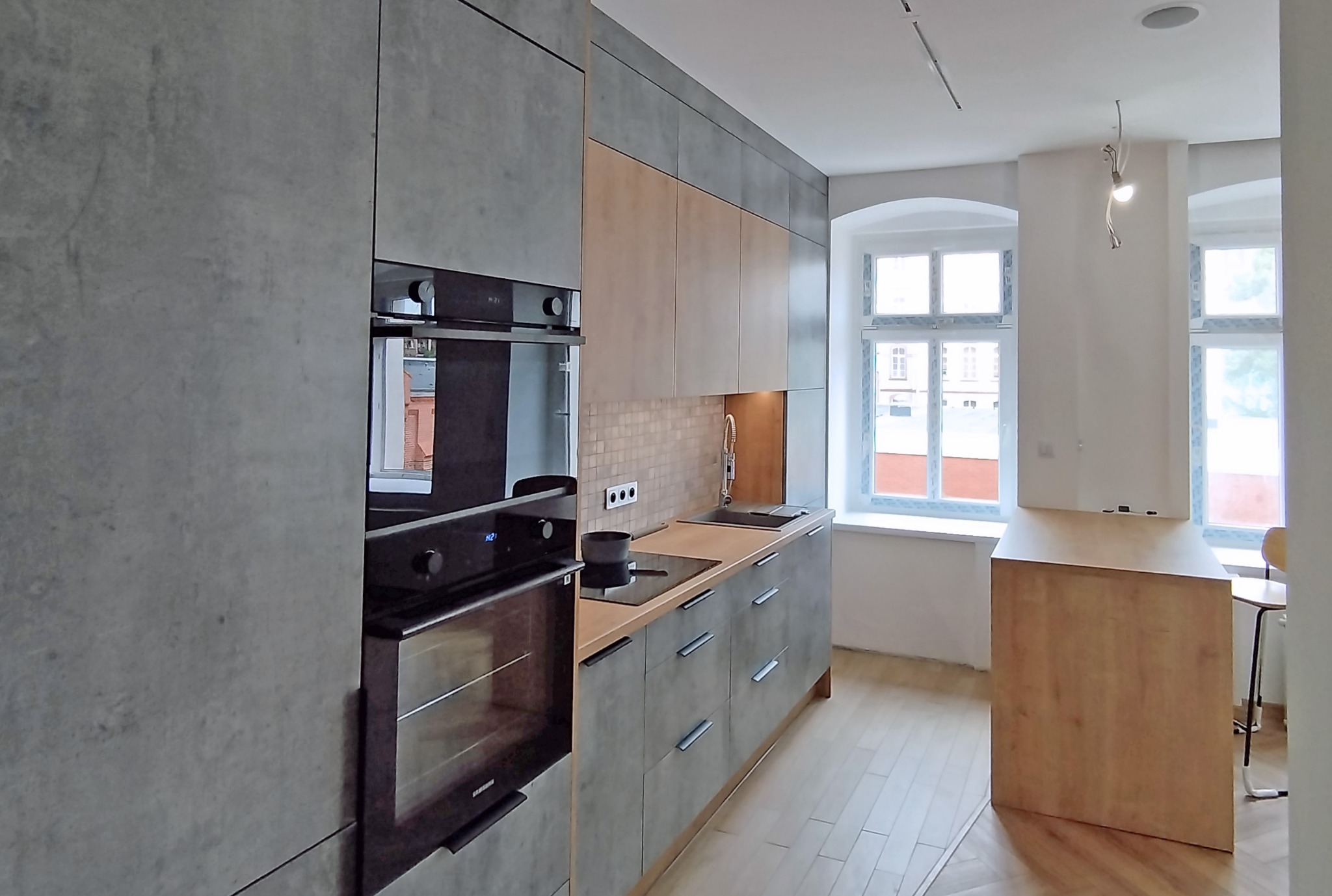 Nowoczesna kuchnia z szarymi frontami i drewnianymi akcentami, wbudowany piekarnik, płyta indukcyjna i okrągłe okna w tle. Minimalistyczny design.