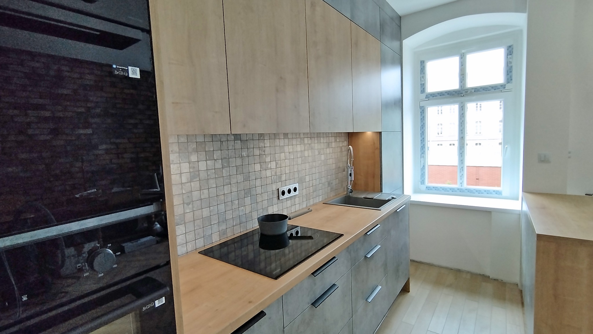 Nowoczesna kuchnia z drewnianymi blatami i szarymi frontami szafek, mozaika na ścianie, okno z widokiem na budynek.