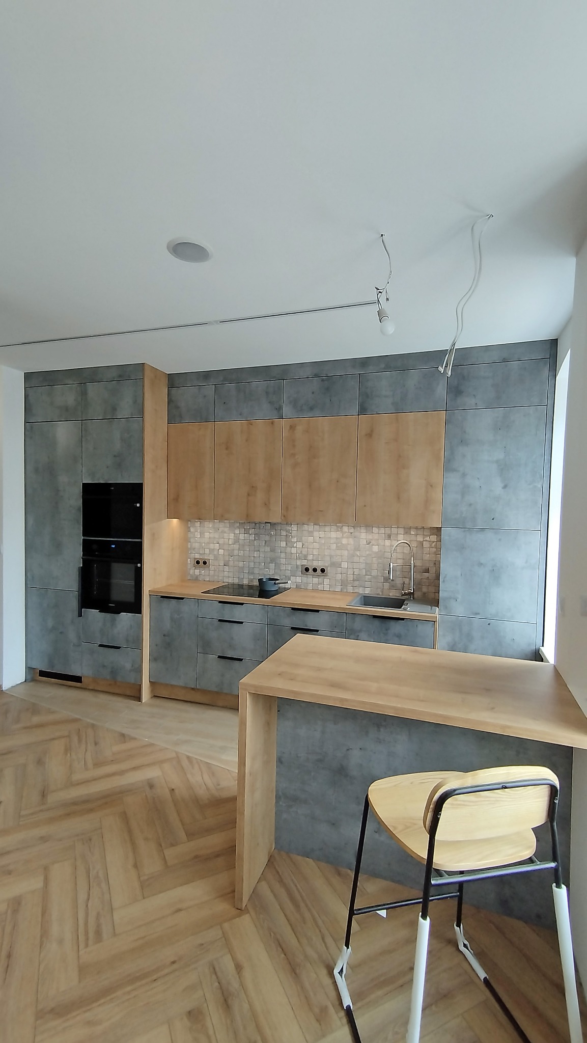 Nowoczesna kuchnia z szarymi frontami imitującymi beton, drewnianymi akcentami i mozaiką nad blatem. Krzesło barowe z jasnego drewna. Parkiet ułożony w jodełkę.