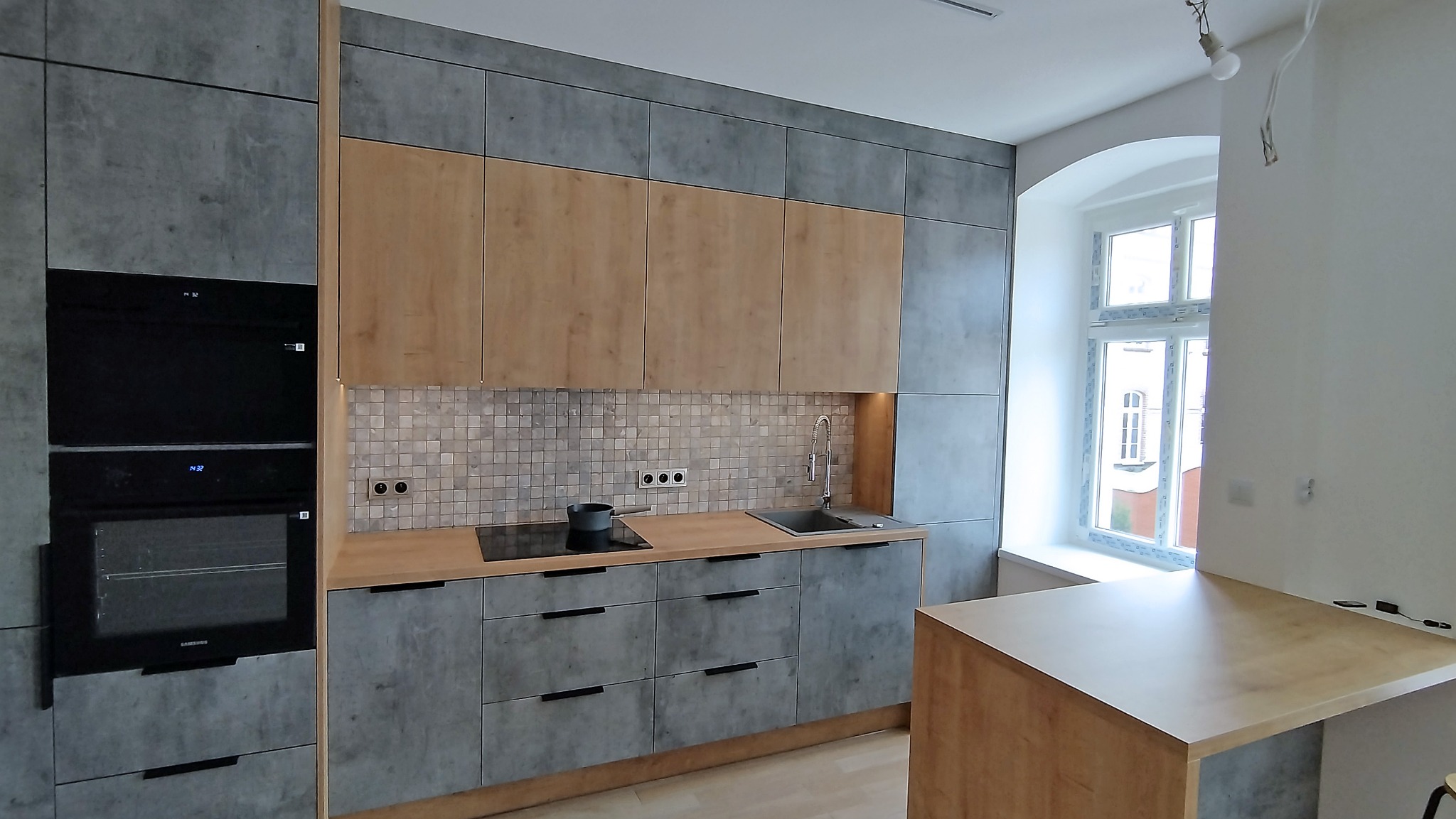 Nowoczesna kuchnia z szarymi frontami i drewnianymi szafkami wiszącymi, mozaika nad blatem, czarna płyta indukcyjna i piekarnik w zabudowie, okno z łukiem.