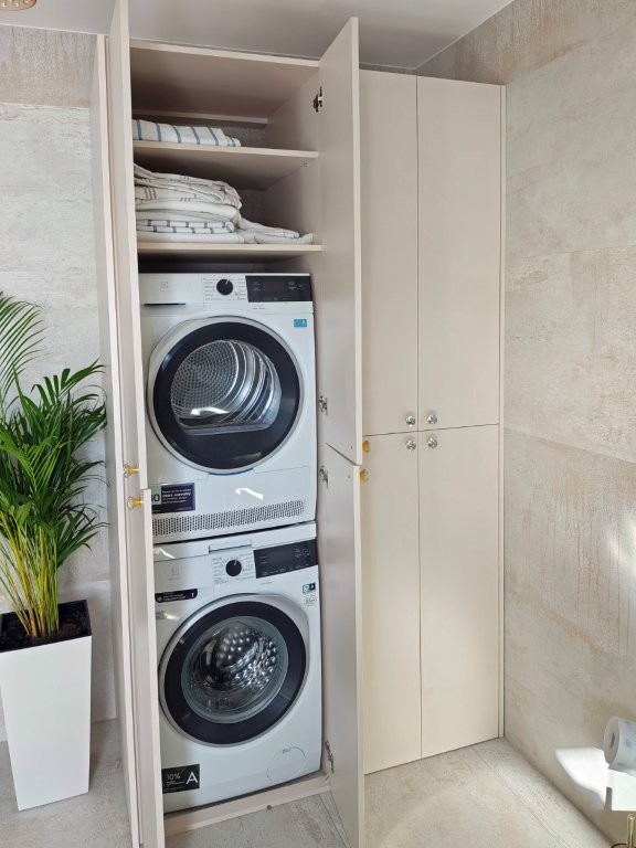 Biała szafa wnękowa z pralką i suszarką w łazience, z półkami na tekstylia. Minimalistyczny design, jasne wnętrze, roślina w doniczce obok.