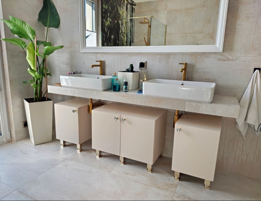 Nowoczesna łazienka z beżowymi szafkami pod umywalki, złotymi bateriami i nogami szafek. Umywalki prostokątne, białe. Na blacie akcesoria i roślina doniczkowa.