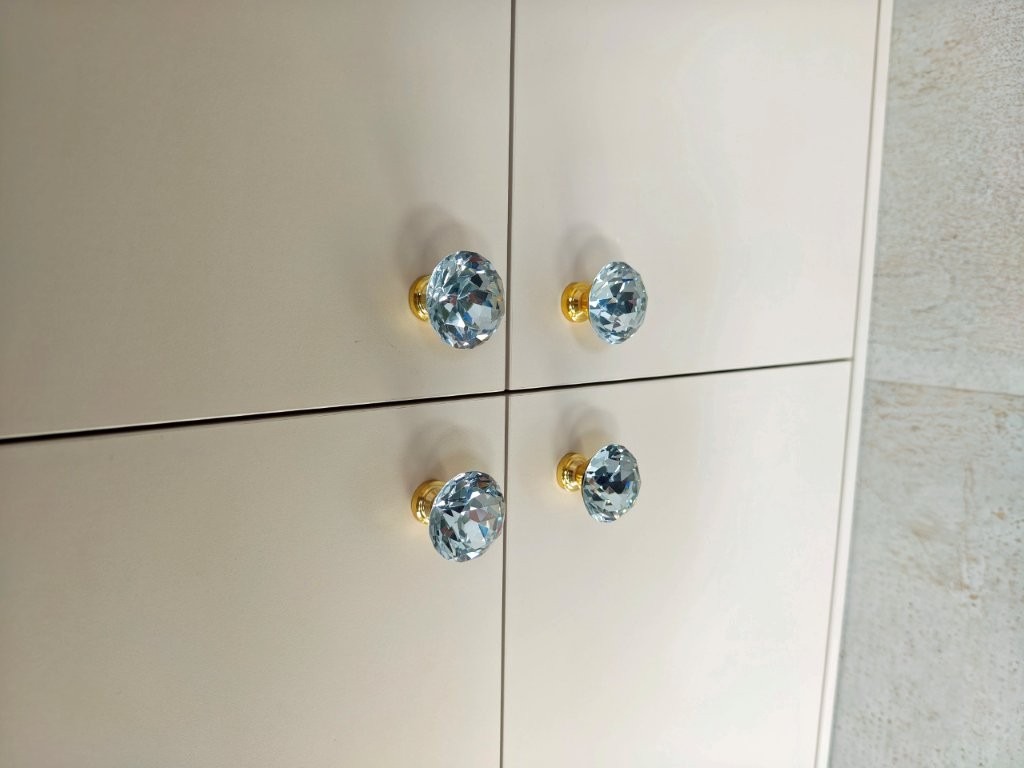 Beżowe fronty szafy z czterema kryształowymi gałkami z pozłacaną podstawą. Minimalistyczny design, subtelne detale, eleganckie wykończenie.