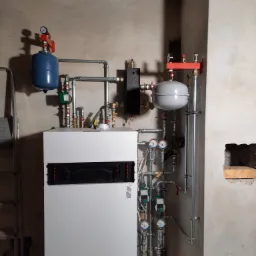 Nowoczesny piec centralnego ogrzewania, z niebieskim i szarym zbiornikiem wyrównawczym, zainstalowany w surowym pomieszczeniu z nieotynkowanymi ścianami, widoczna drabina.