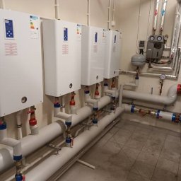 PREZO - Rząd białych pieców centralnego ogrzewania zamontowanych na ścianie, z widocznymi rurami i zaworami, w pomieszczeniu technicznym z szarą podłogą.