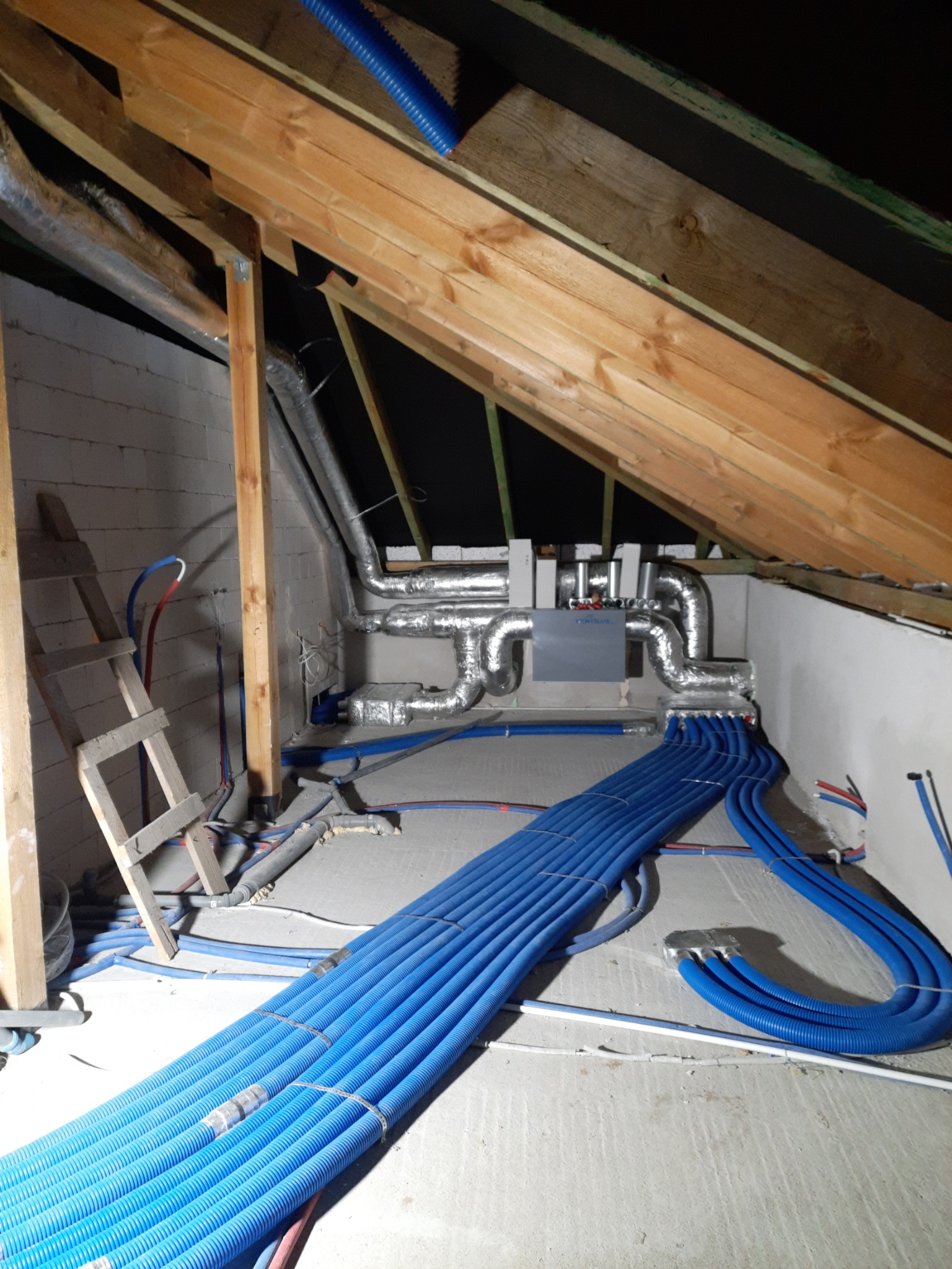 Instalacja wentylacyjna z centralą rekuperacyjną w trakcie montażu na poddaszu, widoczne rury wentylacyjne aluminiowe i niebieskie rury doprowadzające powietrze, drewniana konstrukcja dachu...