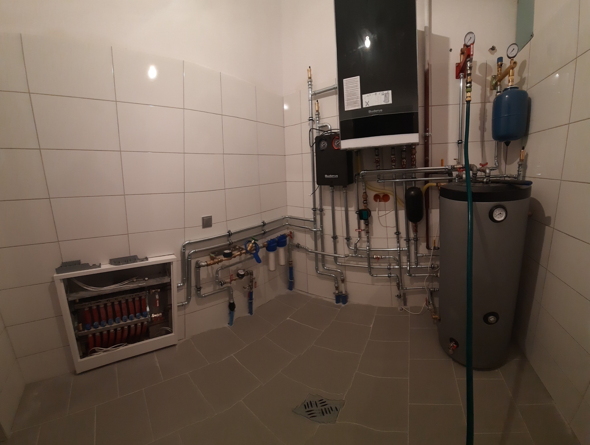 Kotłownia z piecem kondensacyjnym, zasobnikiem ciepłej wody, rozdzielaczem ogrzewania podłogowego i licznymi rurami instalacji grzewczej na tle wykafelkowanych ścian.