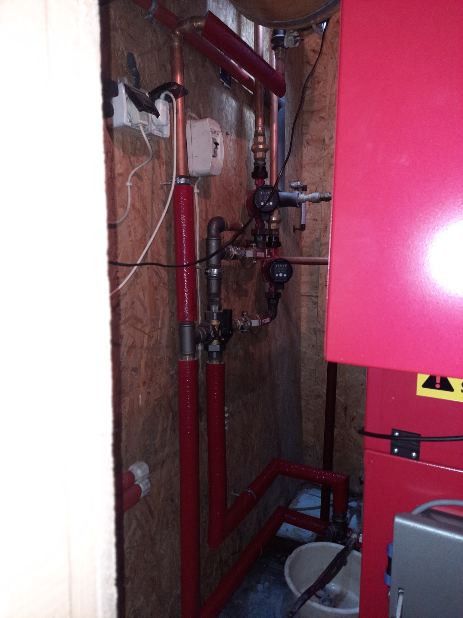 Wnętrze kotłowni z czerwonymi rurami instalacji grzewczej, widoczne pompy obiegowe, osprzęt oraz fragment kotła, całość na tle surowej płyty OSB.