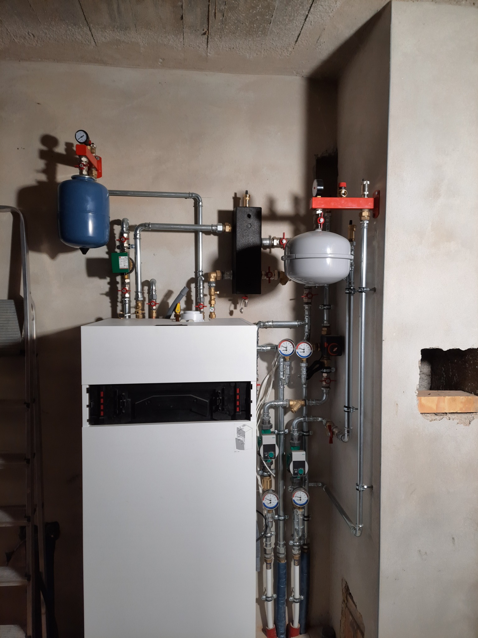 Nowoczesny piec centralnego ogrzewania, z niebieskim i szarym zbiornikiem wyrównawczym, zainstalowany w surowym pomieszczeniu z nieotynkowanymi ścianami, widoczna drabina.