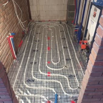 Układanie rur ogrzewania podłogowego na siatce montażowej w surowym wnętrzu łazienki, widoczne przyłącza wodne i fragment ściany z cegły.