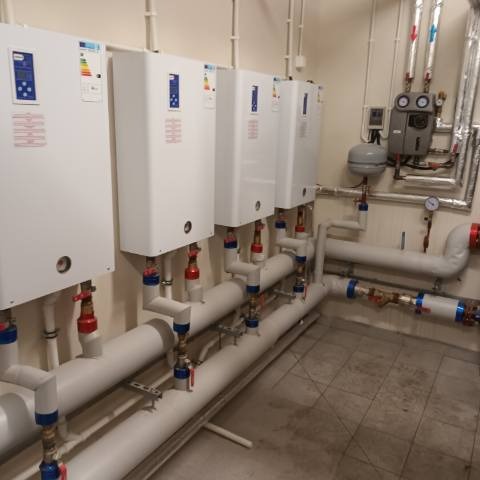 Rząd białych pieców centralnego ogrzewania zamontowanych na ścianie, z widocznymi rurami i zaworami, w pomieszczeniu technicznym z szarą podłogą.