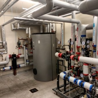 W kotłowni widoczny szary, pionowy zbiornik akumulacyjny ciepła otoczony siecią rur grzewczych z licznymi zaworami, manometrami i izolacją termiczną. Rury pomalowane w różne kolory, niektóre...