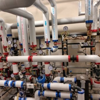 Rozbudowana instalacja grzewcza w kotłowni z licznymi rurami, zaworami, manometrami i czujnikami, oznaczona strzałkami kierunku przepływu, widoczna w pomieszczeniu technicznym.