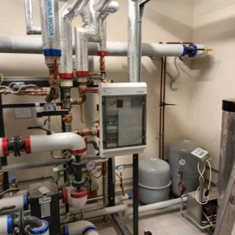 Węzeł cieplny z licznymi rurami w izolacji, manometrami i zaworami. Widoczna szafka sterownicza z elektroniką, zbiorniki wyrównawcze i elementy instalacji grzewczej w kotłowni.