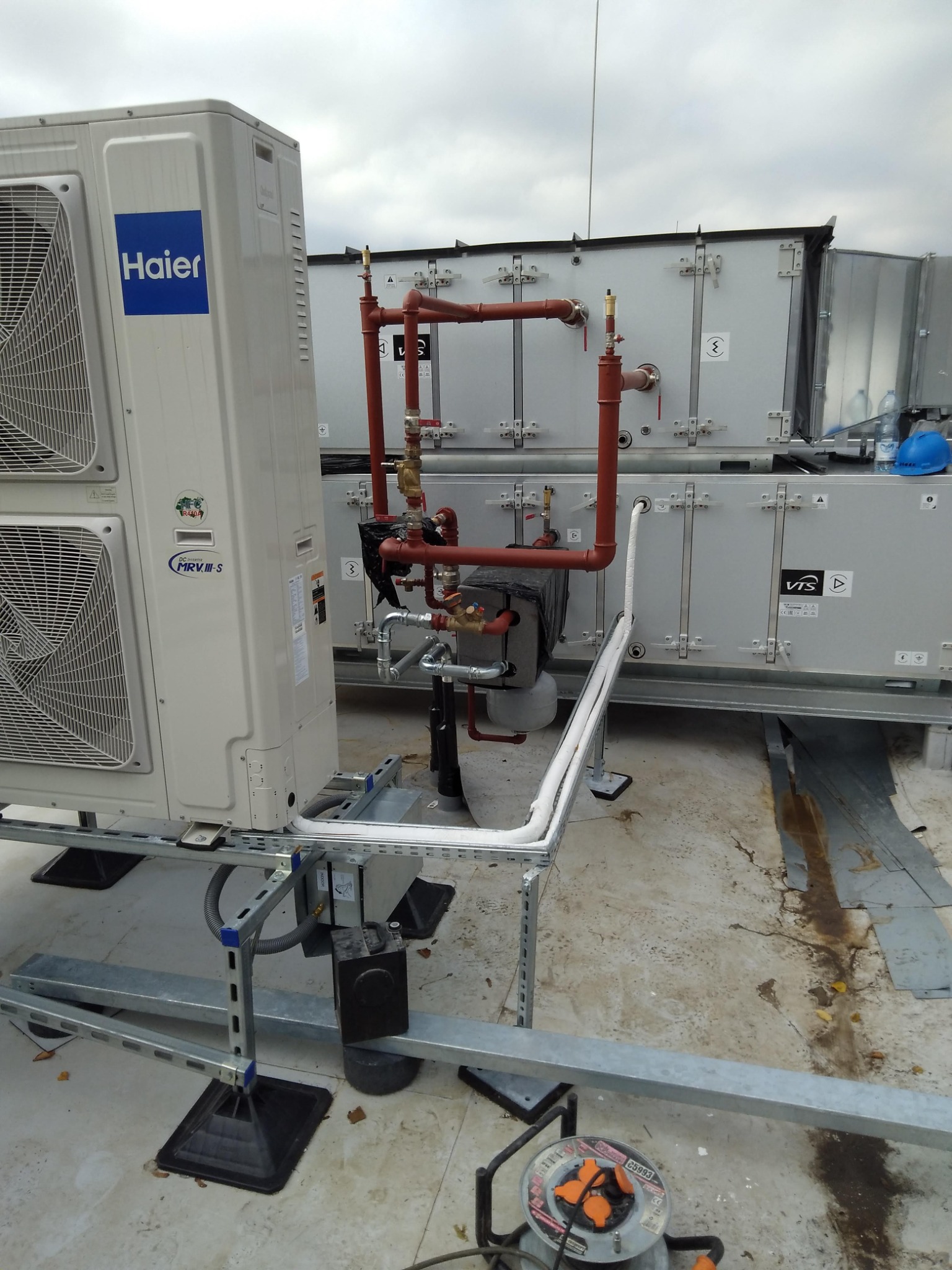 Instalacja klimatyzacji Haier na dachu budynku z widocznymi rurami miedzianymi i izolowanymi przewodami, oraz skrzyniami wentylacyjnymi VTS w tle, z rozłożonym przedłużaczem elektrycznym na płaskim...