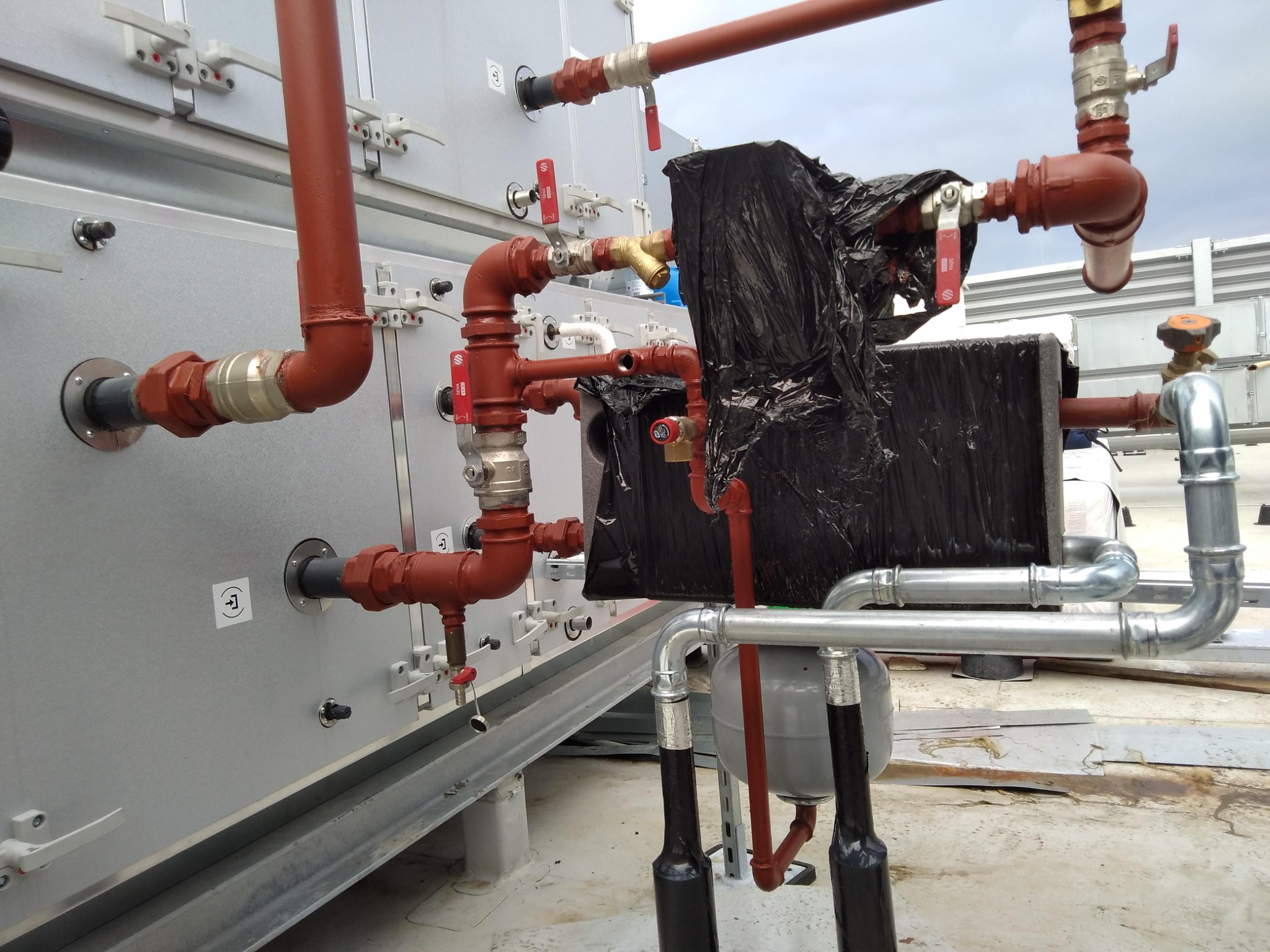 Złożony system rur grzewczych i hydraulicznych z zaworami, częściowo zaizolowany czarną folią, zamontowany na srebrnej obudowie urządzenia na dachu.