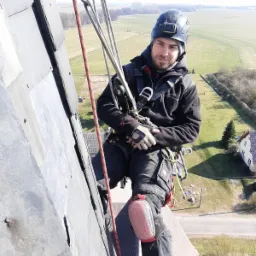 Pracownik w uprzęży, z kaskiem na głowie, zwisa z dachu pokrytego szarą dachówką łupkową, w tle rozległy krajobraz.