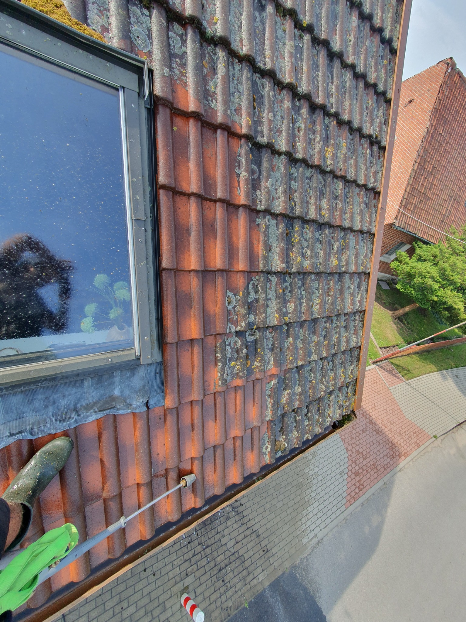 Częściowo oczyszczony dach z czerwonej dachówki obok okna, widoczna lanca ciśnieniowa i fragment buta gumowego, na dachu widoczne porosty i mech.